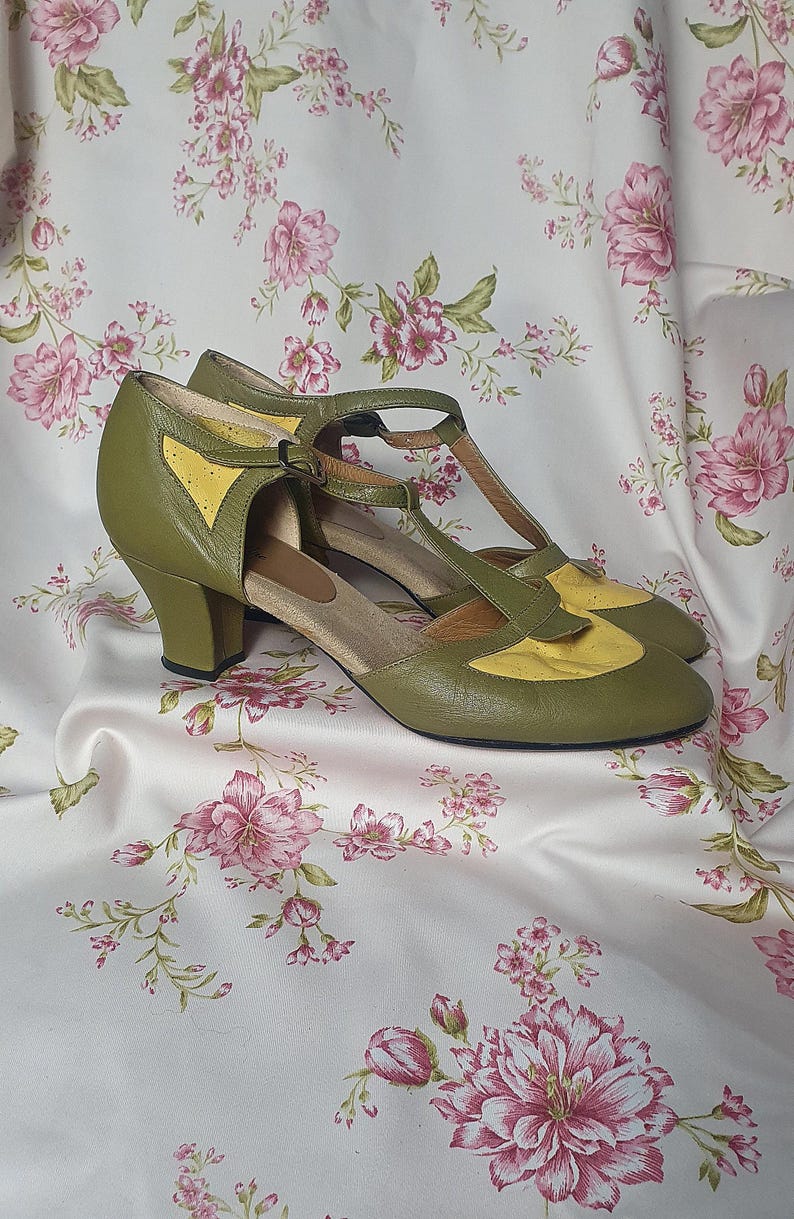 Op de afbeelding: Olijfgroene en gele vintage schoenen met een T-band ontwerp. De schoenen hebben een lage hak en een contrasterend geel paneel op de neus. Ze worden getoond op een stof met bloemenpatroon.