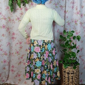 May include: A person wearing a pale yellow knit sweater and a floral print skirt. The skirt features a black background with large, colorful flowers in shades of pink, blue, and purple. The outfit is vintage-inspired.
