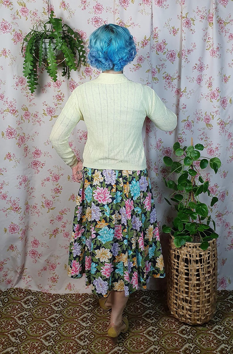 May include: A person wearing a pale yellow knit sweater and a floral print skirt. The skirt features a black background with large, colorful flowers in shades of pink, blue, and purple. The outfit is vintage-inspired.