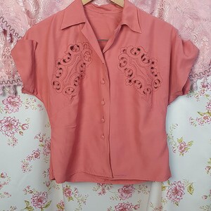 May include: A coral-colored short-sleeve button-down shirt featuring a pointed collar and intricate cut-out embroidery detailing on both sides of the chest. The garment is displayed on a wooden hanger, showcasing a solid color and button closure.