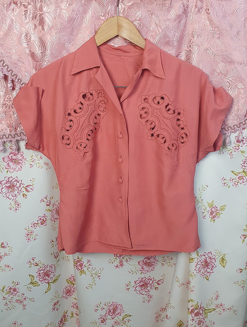 May include: A coral-colored short-sleeve button-down shirt featuring a pointed collar and intricate cut-out embroidery detailing on both sides of the chest. The garment is displayed on a wooden hanger, showcasing a solid color and button closure.