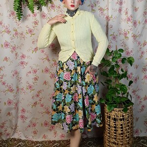 May include: A light yellow cardigan with a scalloped edge is buttoned over a floral print skirt. The skirt features a black background with large pink, blue, and yellow flowers. The outfit is paired with tan shoes.