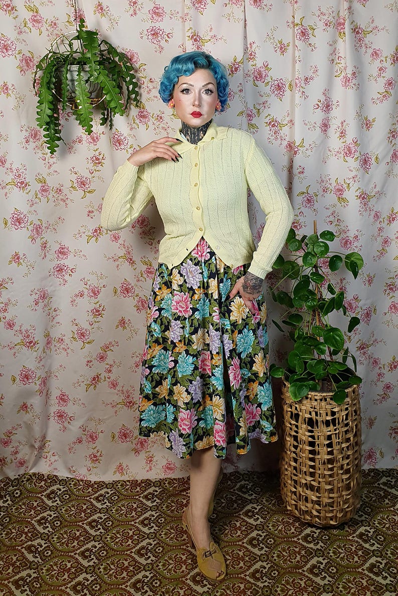 May include: A light yellow cardigan with a scalloped edge is buttoned over a floral print skirt. The skirt features a black background with large pink, blue, and yellow flowers. The outfit is paired with tan shoes.