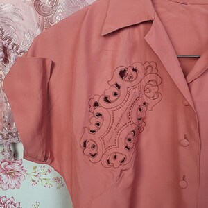 May include: A dusty rose button-down shirt featuring a pointed collar, short cuffed sleeves, and a prominent decorative panel on the chest. The panel displays intricate cutwork embroidery in a matching dusty rose thread, creating a detailed pattern.