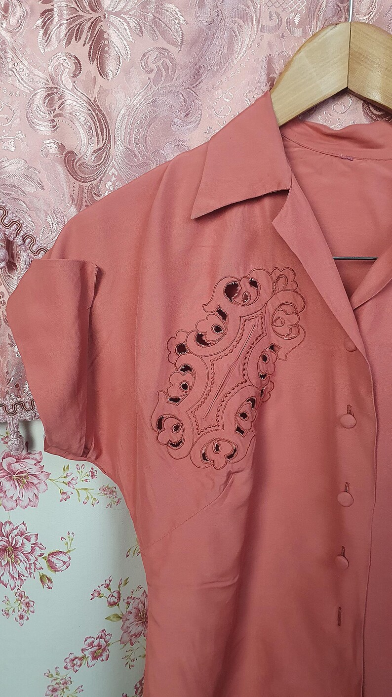 May include: A dusty rose button-down shirt featuring a pointed collar, short cuffed sleeves, and a prominent decorative panel on the chest. The panel displays intricate cutwork embroidery in a matching dusty rose thread, creating a detailed pattern.
