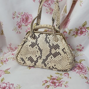 Könnte beinhalten: Eine kleine, cremefarbene Handtasche mit Schlangenhautmuster und goldfarbenen Beschlägen. Die Tasche hat einen oberen Henkel und einen Verschluss mit einem Verschluss.