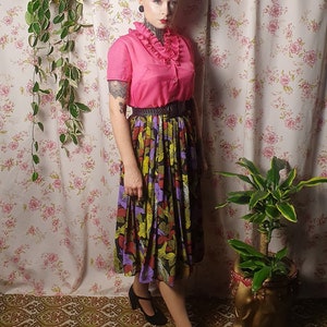 May include: A woman wearing a pink ruffled blouse with a black belt and a colorful floral skirt. She is standing in front of a floral patterned wall with a plant hanging on the left and another plant in a pot on the right.