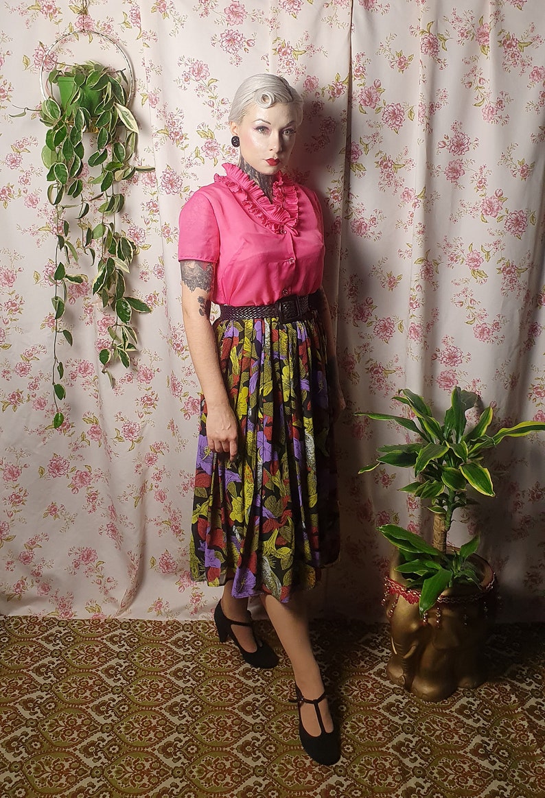 May include: A woman wearing a pink ruffled blouse with a black belt and a colorful floral skirt. She is standing in front of a floral patterned wall with a plant hanging on the left and another plant in a pot on the right.