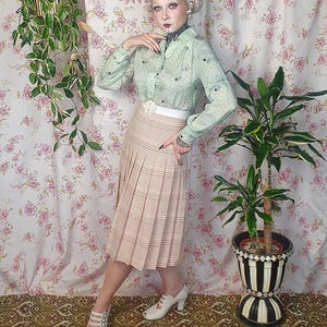 Op de afbeelding: Een lichtgroen overhemd met lange mouwen en knoopsluiting met een subtiel ruitpatroon en een beige geplooide midirok met horizontale strepen. De outfit bevat een witte tailleband met een bloemaccent en witte peep-toe hakken.