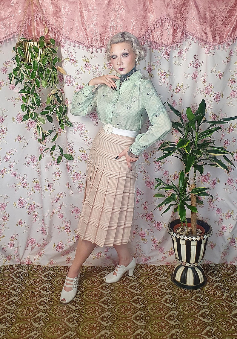 Op de afbeelding: Een lichtgroen overhemd met lange mouwen en knoopsluiting met een subtiel ruitpatroon en een beige geplooide midirok met horizontale strepen. De outfit bevat een witte tailleband met een bloemaccent en witte peep-toe hakken.