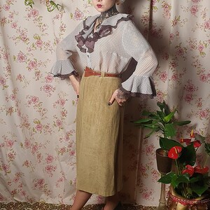 May include: A woman wearing a white and brown polka dot blouse with a brown ruffled collar and a brown belt. She is wearing a khaki skirt and green shoes. She is standing in front of a floral wallpaper.