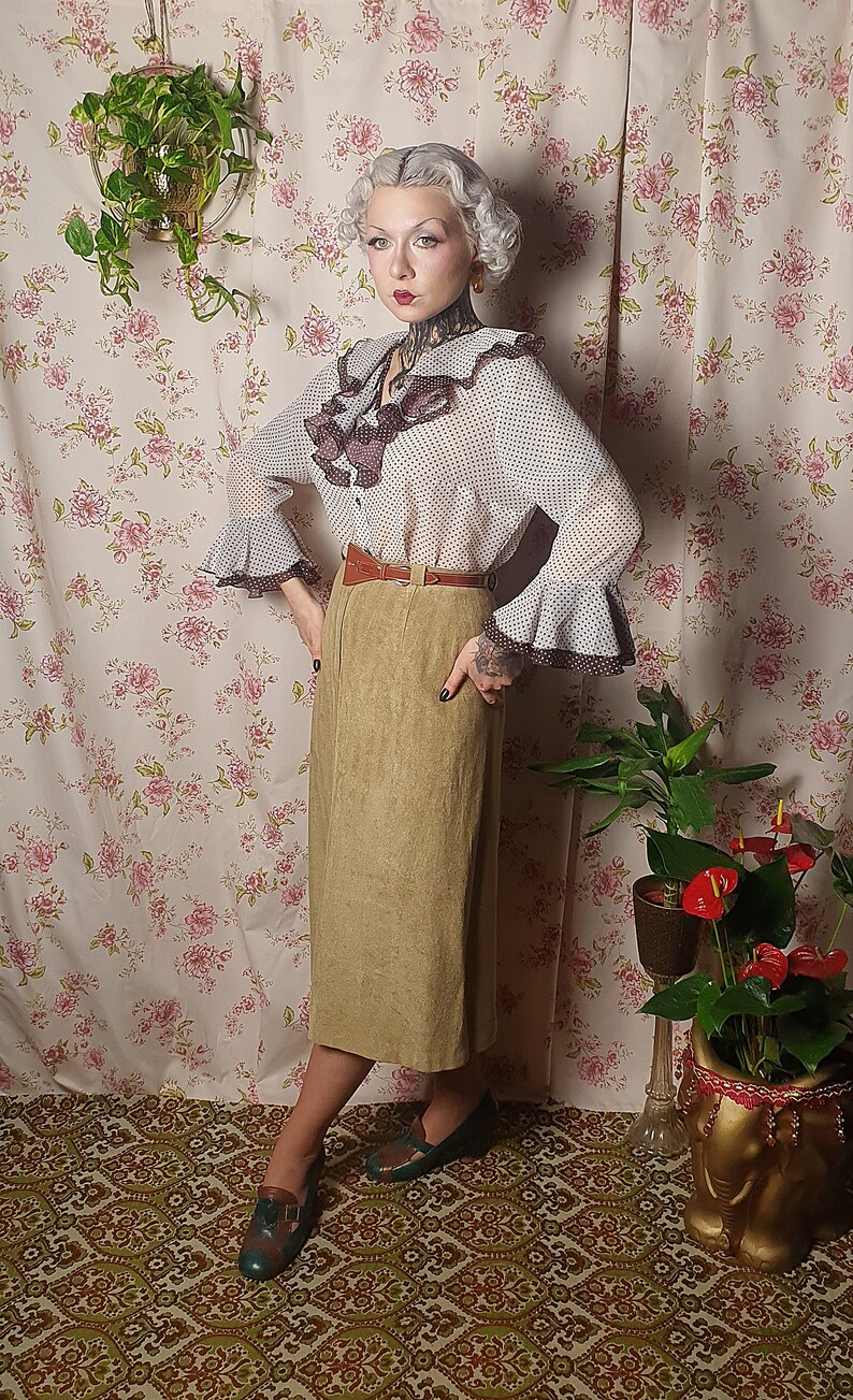 May include: A woman wearing a white and brown polka dot blouse with a brown ruffled collar and a brown belt. She is wearing a khaki skirt and green shoes. She is standing in front of a floral wallpaper.