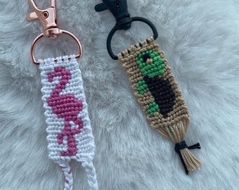 May include: Two macrame keychains with metal lobster clasps. One features a pink flamingo design on a white background with a rose gold clasp. The other displays a green and brown turtle pattern on a tan background with a matte black metal clasp.