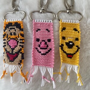 Peut inclure: Trois porte-clés en macramé rectangulaires présentant des visages de personnages tissés. Le porte-clé de gauche est orange et noir avec un nez rose, celui du centre est rose avec des traits noirs, et celui de droite est jaune avec des détails noirs et rouges.