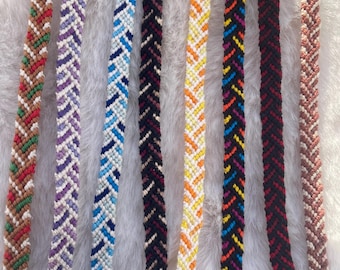 May include: Eight handmade woven friendship bracelets arranged vertically. Each bracelet features a distinct chevron pattern in various color combinations, including brown with green and red, purple with white, blue with white, black, yellow with orange, black with multi-colored accents, solid black, and light brown.
