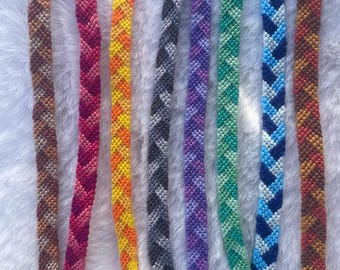 May include: A collection of handmade friendship bracelets in various colors, including brown, red, yellow, gray, purple, green, blue, and orange. Each bracelet features a chevron pattern and a braided end.