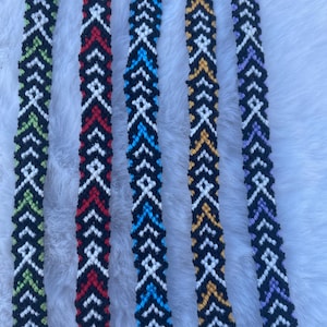 May include: Five handmade friendship bracelets with geometric patterns. The bracelets feature black, white, and various accent colors, including green, red, blue, yellow, and purple. Each bracelet is approximately 0.5 inches wide.