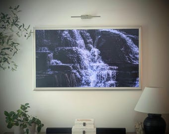 Op de afbeelding: Een ingelijst digitaal kunstwerk met een zwart-wit watervalscène. Het kunstwerk is aan een witte muur bevestigd, met een kleine lamp erboven. Een zwarte lamp met een witte kap en decoratieve planten staan op de voorgrond.