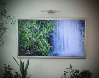Op de afbeelding: Een ingelijste print van een watervalscène met weelderig groen gebladerte aan de linkerkant en trapsgewijs water aan de rechterkant. De print is gemonteerd op een witte muur, met een kleine lamp erboven. Het thema is natuur en landschap.