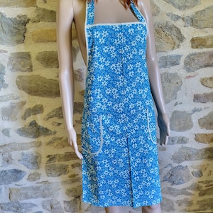 Könnte beinhalten: Eine blaue Schürze mit weißem Blumenmuster und weißer Bordüre. Die Schürze hat zwei Vordertaschen und einen Faltenrock. Die Träger sind weiß und gehen über die Schultern. Die Schürze wird auf einer Schaufensterpuppe präsentiert.