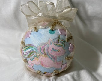 May include: A handmade Christmas ornament featuring a unicorn with a rainbow mane and tail on a blue background. The ornament is decorated with gold glitter and a white satin ribbon.