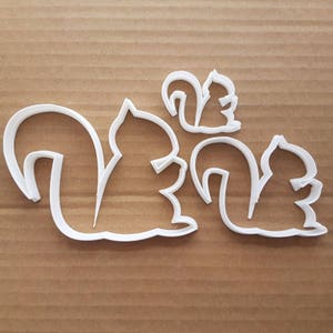 May include: Three white plastic squirrel-shaped cookie cutters of varying sizes. The squirrels have fluffy tails and are in a side profile. The cutters are on a brown cardboard surface. Ideal for baking themed cookies.