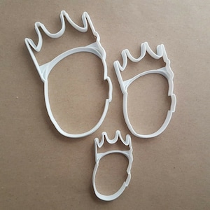 Puede incluir: Tres cortadores de galletas de plástico blanco con forma de cabeza con una corona en la parte superior. Los cortadores son de diferentes tamaños.