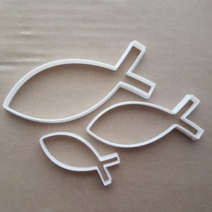 May include: Three white plastic cookie cutters shaped like fish. The cutters are different sizes, with the largest cutter at the top of the image and the smallest cutter at the bottom.