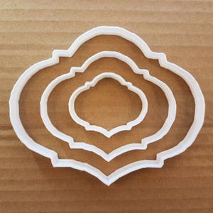 以下が含まれることがあります： 装飾的なフレームの形をした白いプラスチック製のクッキーカッターが3つ。カッターは互いにネストされており、最も小さいカッターが中央にあります。