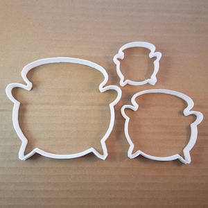 May include: Three white plastic cookie cutters shaped like cauldrons. The cutters are different sizes, with the largest cutter in the centre and the smallest cutter at the top.