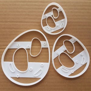 May include: Three white plastic cookie cutters shaped like eggs with different patterns of circles and lines.