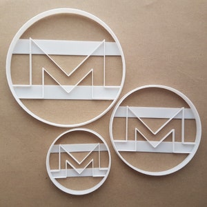May include: Three white plastic cookie cutters in the shape of the letter "M" with a horizontal line across the middle. The cutters are in different sizes.