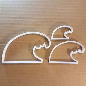 May include: Three white plastic wave-shaped cookie cutters of varying sizes. The cutters have a curved top and a scalloped edge to represent the crest of a wave. The cutters are arranged on a brown surface.