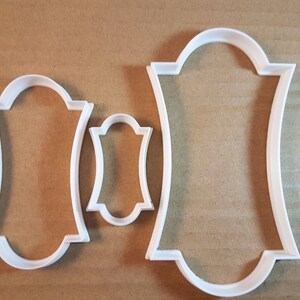 May include: Three white plastic cookie cutters shaped like frames. The largest frame is about 10 cm tall and 7.5 cm wide. The smallest frame is about 3.75 cm tall and 2.5 cm wide.