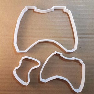 Peut inclure: Deux emportes-pièces en plastique blanc en forme de manettes de jeu vidéo. Le plus grand emporte-pièce est une manette standard avec deux boutons et un pavé directionnel. Le plus petit emporte-pièce est une manette plus moderne avec deux joysticks et quatre boutons.
