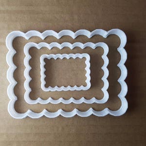 Puede incluir: Tres cortadores de galletas blancos con bordes festoneados en tamaños graduados, anidados entre sí. Los cortadores son rectangulares con un borde ondulado, perfectos para crear galletas o pasteles decorativos. Los cortadores son de plástico.