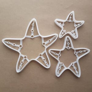 May include: Three white plastic cookie cutters shaped like starfish. The largest cutter is about 6 inches wide and has a detailed design with small circles around the edges. The other two cutters are smaller and have a simpler design.
