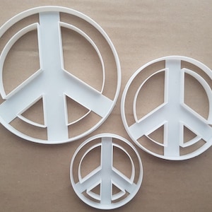 May include: Three white plastic peace sign cookie cutters of different sizes. The largest cutter is about 12.5 cm in diameter.