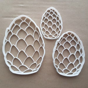 May include: Three white plastic cookie cutters shaped like pine cones. The cutters are different sizes, with the largest cutter in the center of the image.