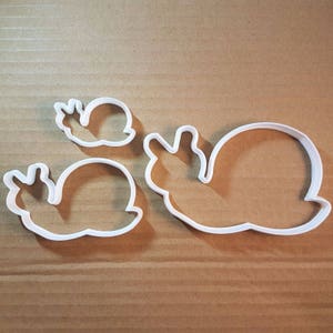 Puede incluir: Tres cortadores de galletas de plástico blanco con forma de caracoles. El caracol más grande está en el centro de la imagen, con un caracol más pequeño a la izquierda y un caracol diminuto en la parte superior derecha.