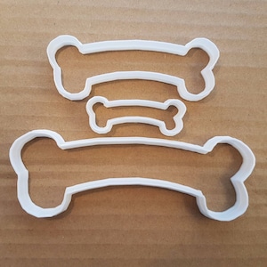 Puede incluir: Tres cortadores de galletas de hueso de perro de plástico blanco de diferentes tamaños. El hueso más grande es curvo, mientras que los dos huesos más pequeños son rectos.