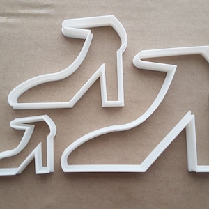 May include: Three white plastic cookie cutters shaped like high-heeled shoes. The cutters are arranged in a row, with the largest cutter at the back and the smallest cutter at the front.