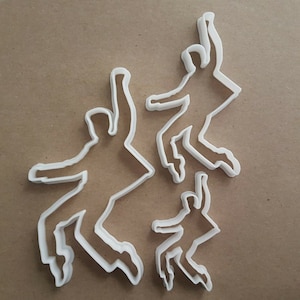 Pode incluir: Três cortadores de biscoitos de plástico branco em forma de figuras dançando. As figuras estão em poses diferentes, com uma figura no ar com os dois braços levantados, uma figura com um braço levantado e um braço para baixo, e uma figura com os dois braços para baixo.
