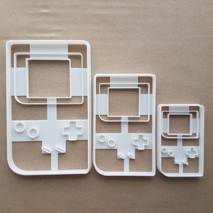 May include: Three white plastic cookie cutters shaped like a Game Boy handheld video game console. The cutters are in different sizes, with the largest cutter on the left and the smallest cutter on the right.