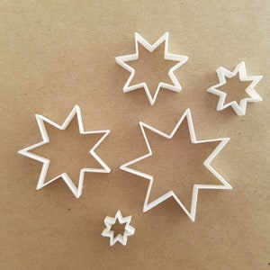 May include: Set of five white plastic cookie cutters shaped like eight-pointed stars. The cutters are arranged on a brown surface.