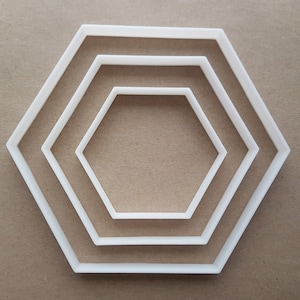 May include: Three white plastic hexagon cookie cutters of different sizes. The cutters are arranged in a concentric pattern, with the largest cutter on the outside and the smallest cutter on the inside.