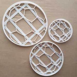 Puede incluir: Tres cortadores de galletas de plástico blanco de diferentes tamaños. Cada cortador tiene una forma circular con un patrón geométrico de hexágonos y líneas diagonales. Están colocados sobre una superficie marrón.