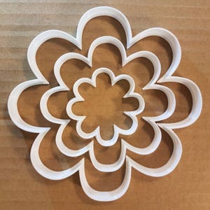 Puede incluir: Cortador de galletas de plástico blanco en forma de flor con cuatro capas de pétalos. El cortador está diseñado para crear una forma de flor en capas para galletas, fondant u otras artesanías comestibles.