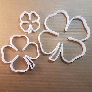 May include: Three white plastic four-leaf clover cookie cutters of different sizes. The largest cutter is in the shape of a four-leaf clover with a stem.
