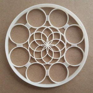 May include: White plastic cookie cutter in the shape of a circle with a geometric design in the center. The design features a flower with multiple layers of petals.